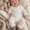Lachende baby in een boxpakje met konijntje print naast een knuffelkonijn, liggend op een zacht bed